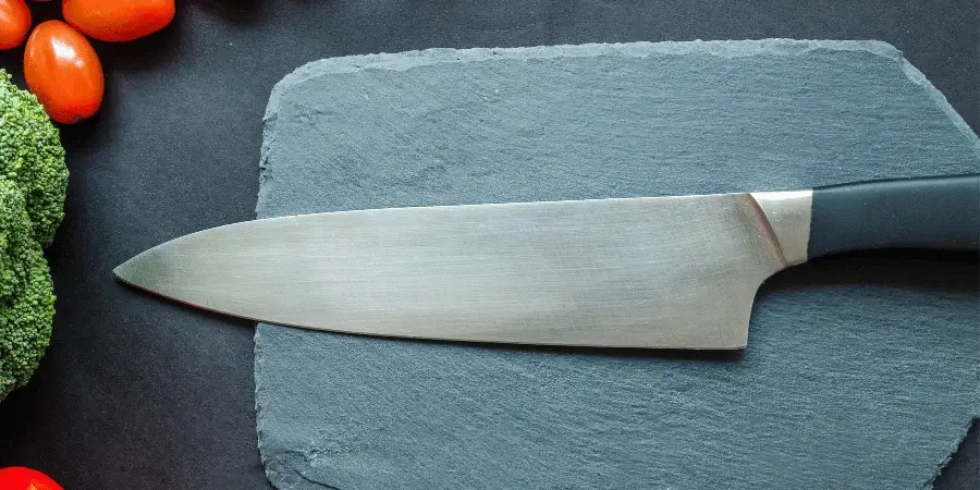 A kitchen knife placed on a slate surface next to fresh vegetables.