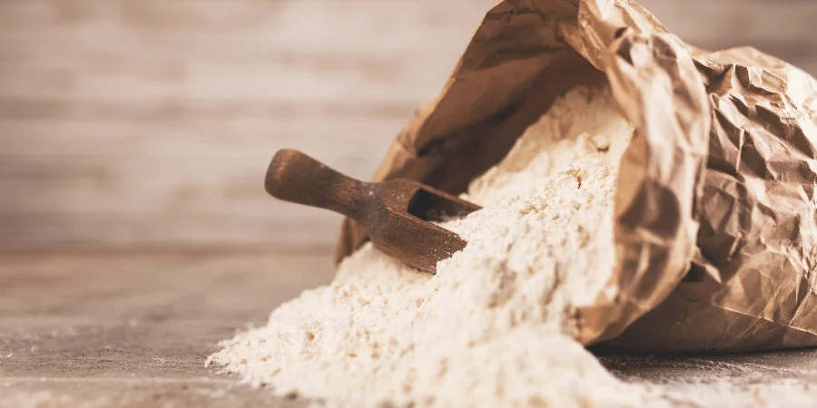 A paper sack filled with flour, with a wooden scoop lying beside it, showing its texture and fine powder.