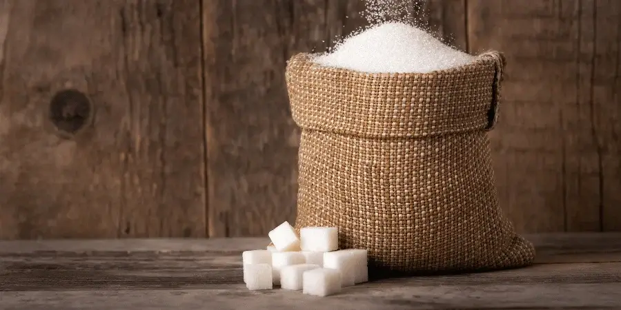 A jute sack filled with white sugar, with sugar cubes scattered beside it, placed on a rustic wooden surface.