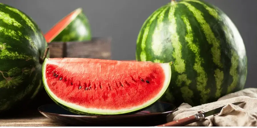 A sliced watermelon on a plate with two whole watermelons in the background.