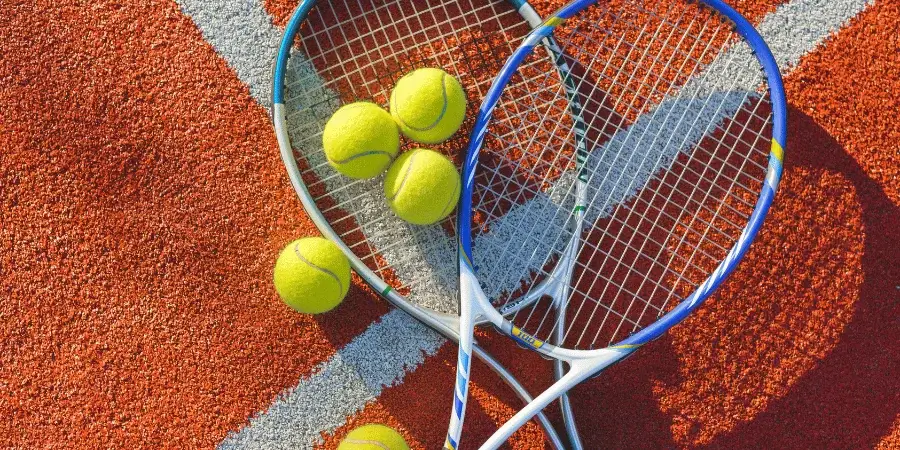 Two tennis rackets with several yellow tennis balls on an orange clay tennis court.