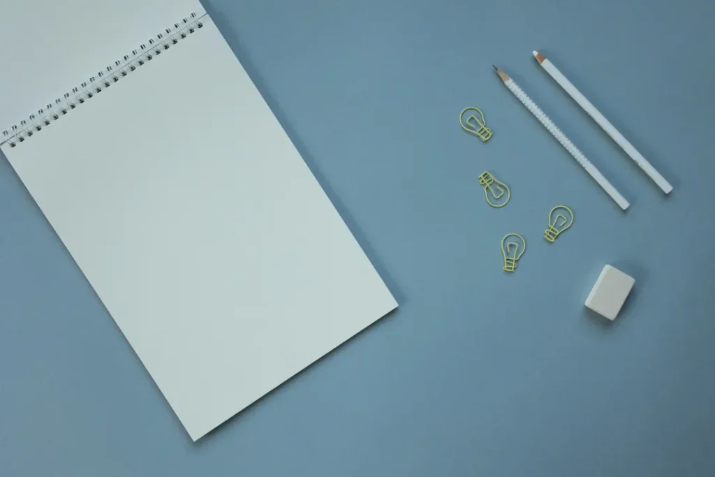 Flat lay of blank spiral notebook, pencils, eraser, and lightbulb paper clips on blue desk.