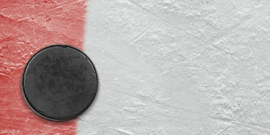 Black hockey puck resting on ice rink surface near red line, surrounded by textured ice.