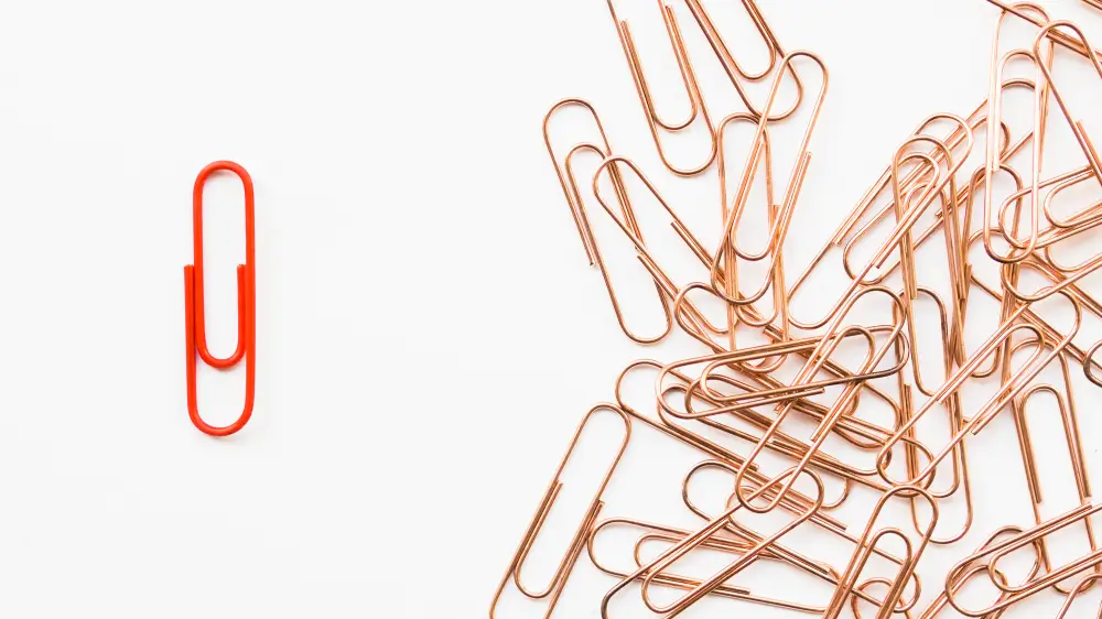 One red paperclip separated from a scattered pile of copper paperclips on a white background.