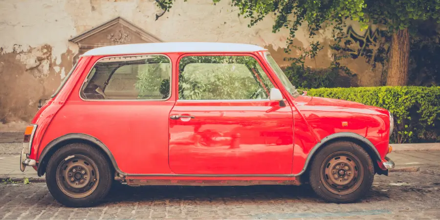 A vintage red small compact car parked on a cobbled street surrounded by greenery and buildings.