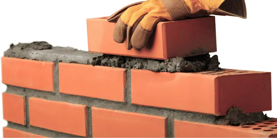 A bricklayer wearing gloves, carefully placing a brick on a newly built brick wall with mortar in between.