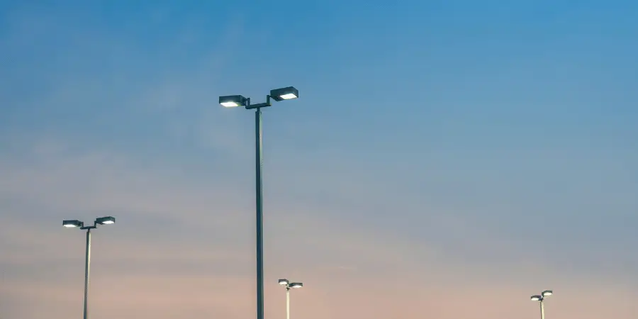 Modern light poles glowing brightly against a twilight sky, illuminating parking lots and outdoor urban spaces.