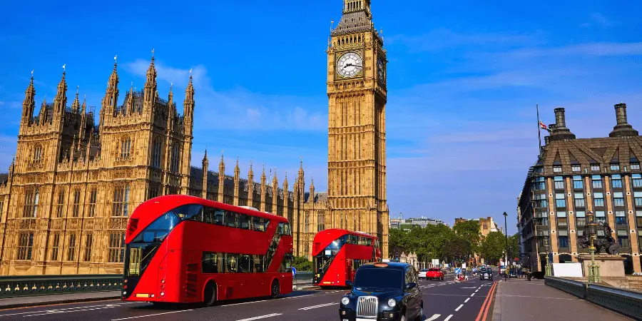 Iconic red double-decker buses drive past Big Ben and the Houses of Parliament in London on a clear sunny day.