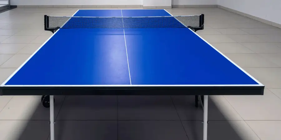 Blue ping pong table with net set up in an indoor room with tiled floor, ready for table tennis match.