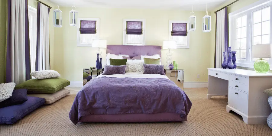 Modern bedroom with purple bedding, green accents, and elegant decor featuring natural light and stylish furniture.