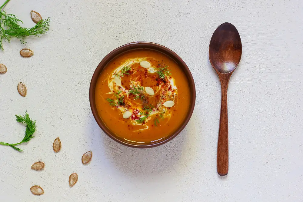 A bowl of creamy pumpkin soup garnished with cream, seeds, and herbs, accompanied by a wooden spoon on a light surface.