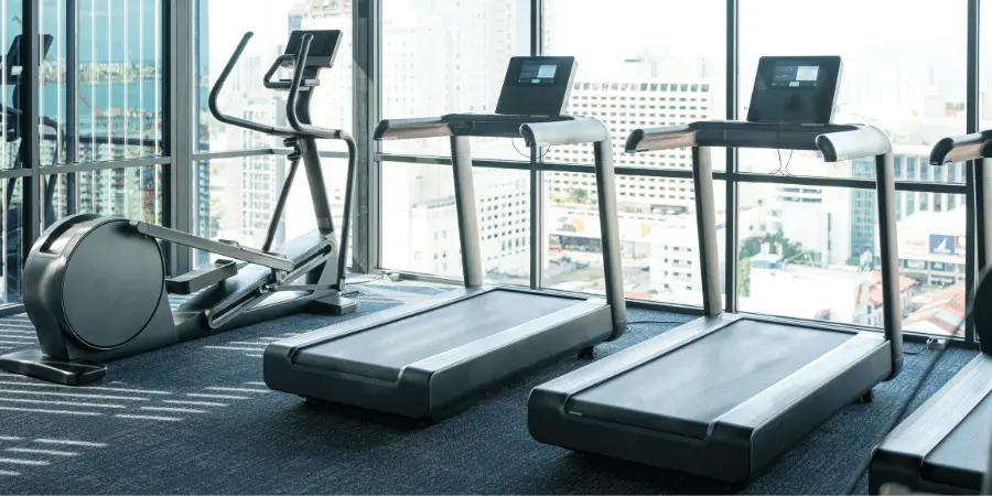 Modern gym with treadmills and elliptical machine facing glass windows overlooking a city skyline view.