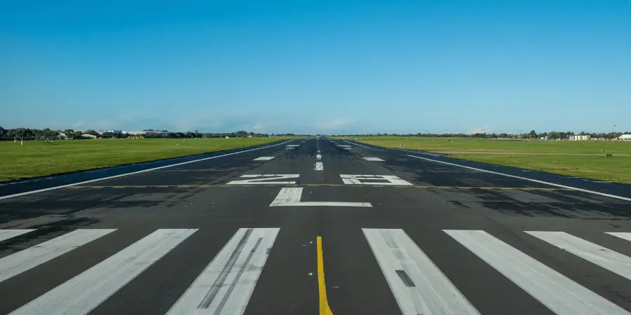 Empty airport runway stretching forward with clear markings, aviation infrastructure under bright blue sky and open green fields.