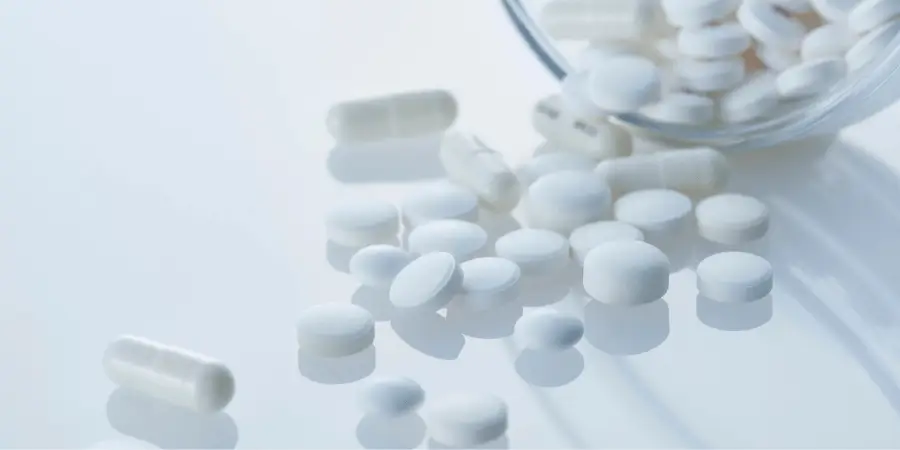 White tablets and capsules scattered on a glossy surface with more pills spilling from a container.