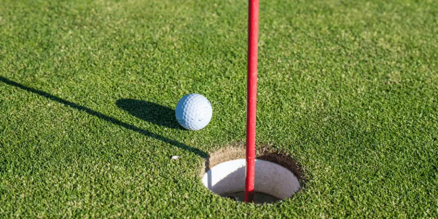 Golf ball resting on the green right beside a golf hole with a red flagstick.