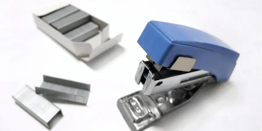 A small blue stapler placed beside an open box of metal staples on a white surface.